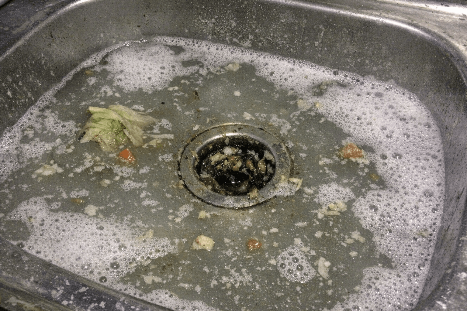 Nahaufnahme eines teilweise mit Speiseresten gefüllten Spülbeckens, das Wasser staut sich am Ablauf